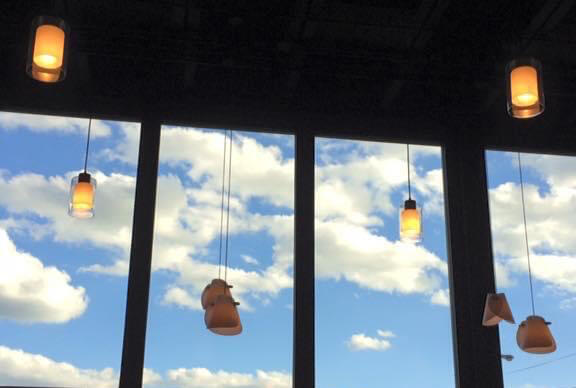 Vertical window panes and vertical hanging lights in front of a brightly lit blue sky with puffy clouds